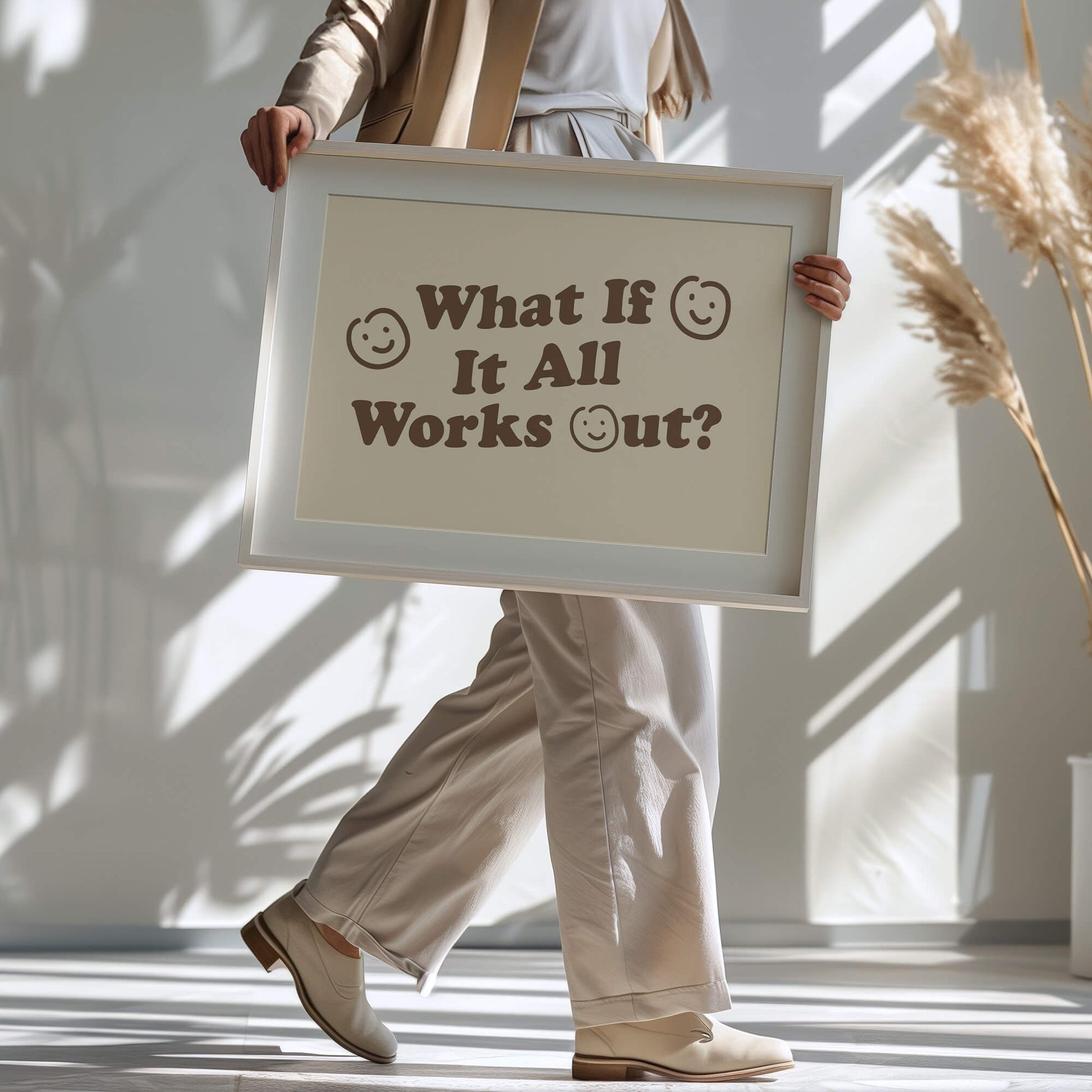 Woman holding a large framed print with a motivational quote reads &quot;what if it all works out?&quot;. The wall art has smiley faces and neutral coloured background with brown lettering