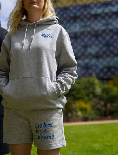 The best is yet to come shorts. positive messaging mental health shorts. matching shorts and hoodie in grey and blue. Stood in park in front of Birmingham Library