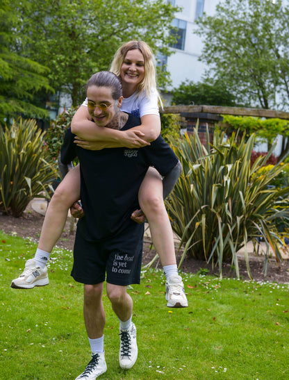 The best is yet to come shorts. positive messaging mental health shorts. Male and female model having piggy back rides in the park