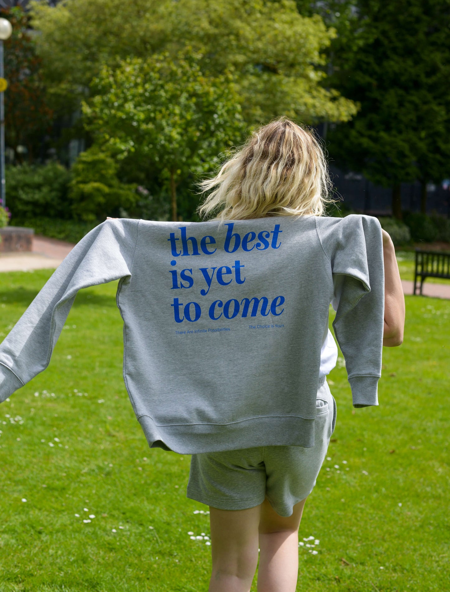 The best is yet to come hoodie. positive messaging mental health hoodie. Girl in nature wearing positive mental health hoodie in grey and blue. Woman walks through park wearing hoodie loosely on shoulders