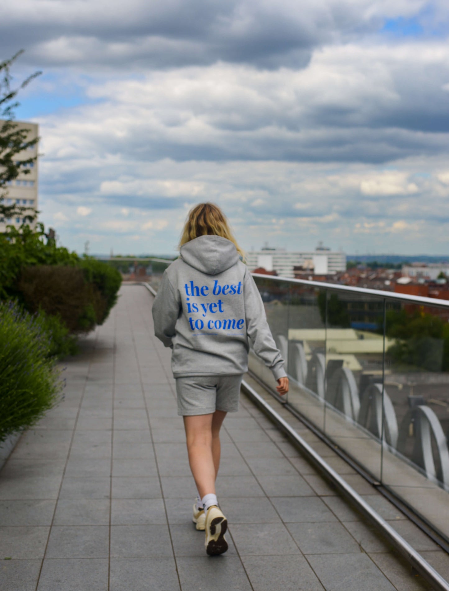 The best is yet to come hoodie in grey. positive messaging mental health hoodie. Woman walks across rooftop wearing affirmation essential hoodie