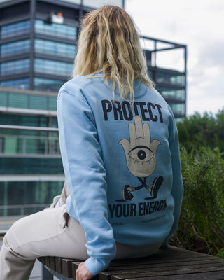 Protect your energy hamsa hand sweater in baby blue. Blonde model wearing positive streetwear. Female model sitting on bench on rooftop