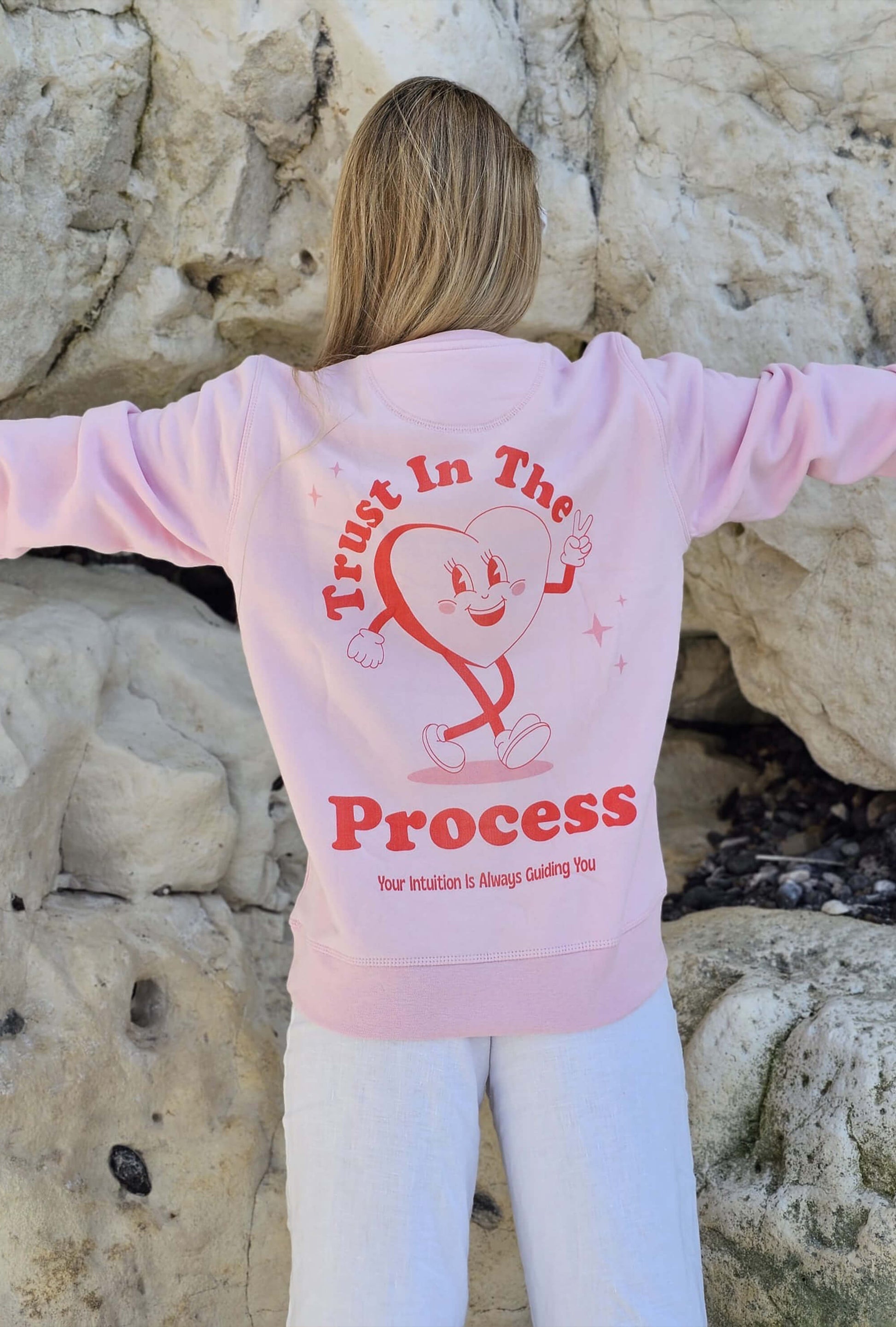 Pink positive sweater with retro mascot that says trust in the process. woman is on the beach
